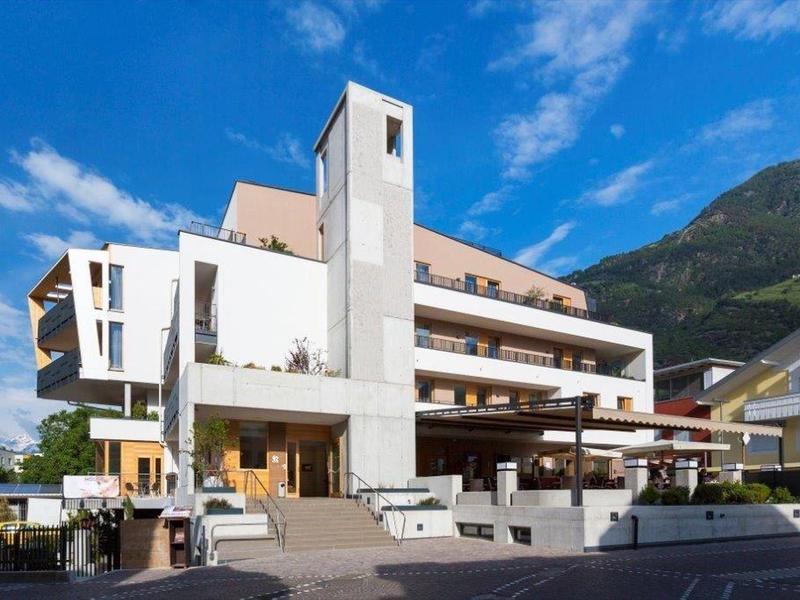Modernes, mehrstöckiges Gebäude mit weißer Fassade vor Bergkulisse und blauem Himmel.