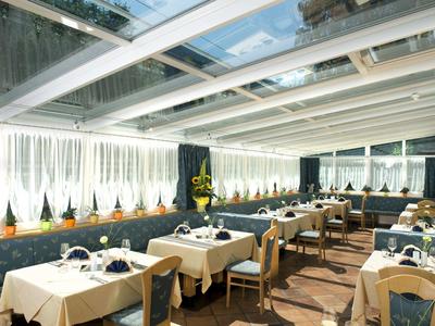 Restaurant mit weißen Tischdecken, blauen Stühlen und großen Fenstern mit weißen Gardinen.