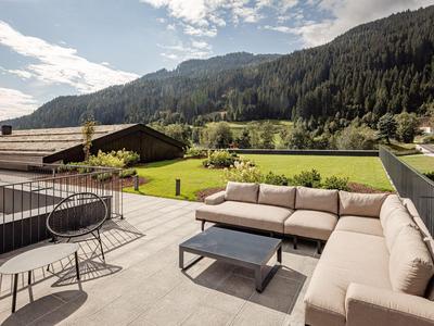 Cozy terrace with sofas and tables overlooking green meadows and forested mountains.