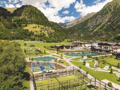 Hotelový komplex v zeleném údolí s bazény, tenisovými kurty a horami v pozadí.
