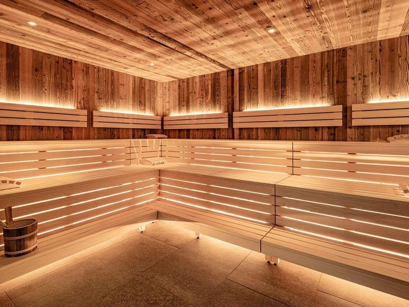 Světlá sauna s dřevěnými lavicemi, teplým osvětlením a dřevěným vědrem na podlaze.