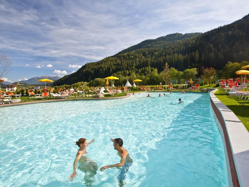 Dvě osoby v venkovním bazénu s lehátky a horami v pozadí během slunečného dne.