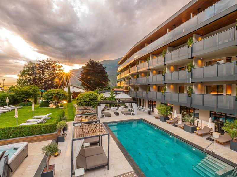 Modern hotel met buitenzwembad, ligstoelen en tuin onder een bewolkte zonsondergang.