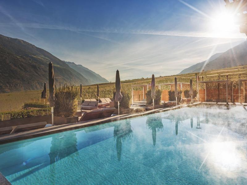 Zonnig buitenzwembad met ligstoelen tegen bergachtergrond onder een heldere hemel.
