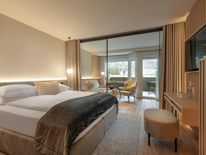 Moderne hotellkamer met groot bed, zithoek, glazen scheidingswand en warm licht.