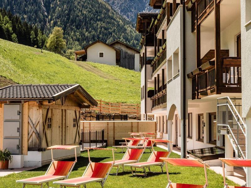 Zonnige ligstoelen voor berghotel met weiden en besneeuwde toppen op de achtergrond.
