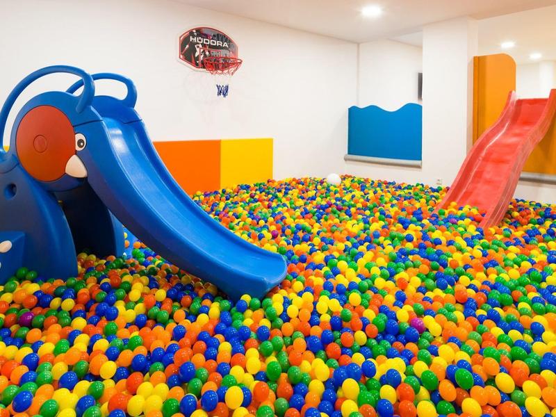 Heldere, kleurrijke ballenbak met twee kleine glijbanen in een binnenruimte.