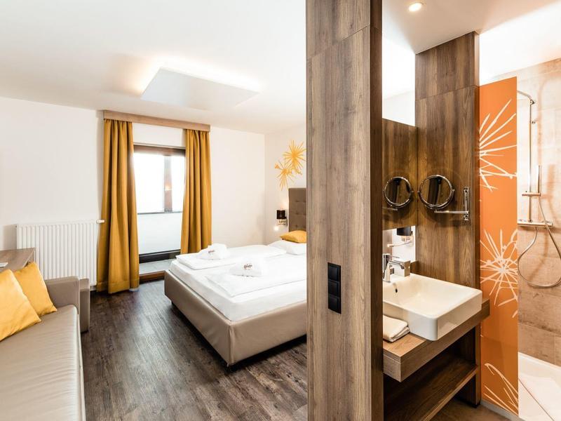 Modern hotelkamer met tweepersoonsbed, sofa, tv en een douche met glazen wand.