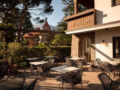 Słoneczny taras z stołami i krzesłami obok budynku hotelowego i zielonego ogrodu.