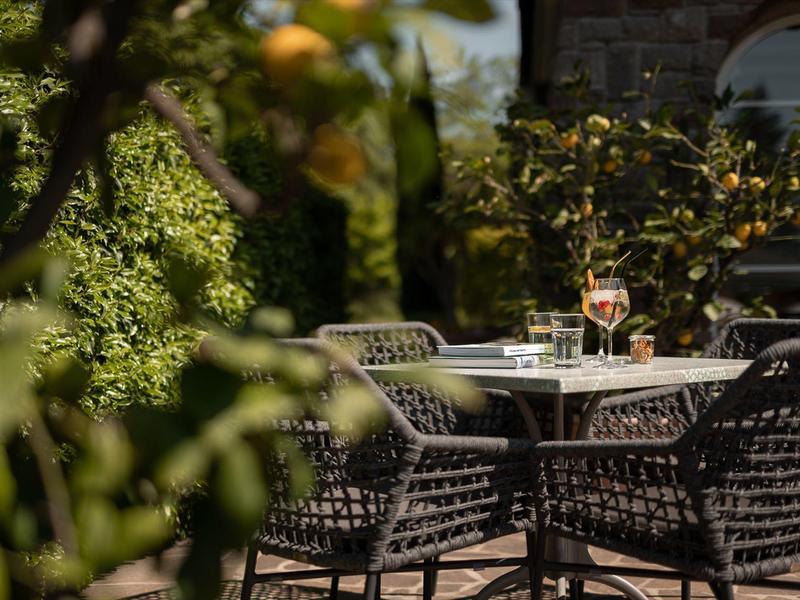 Een rustige terras met een tafel, twee wijnglazen en zwarte stoelen omgeven door groene planten.