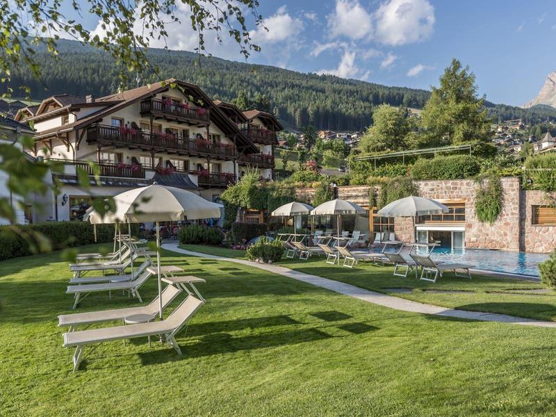 Hotel garden with lounge chairs, umbrellas, pool, and mountains in the background under clear sky.