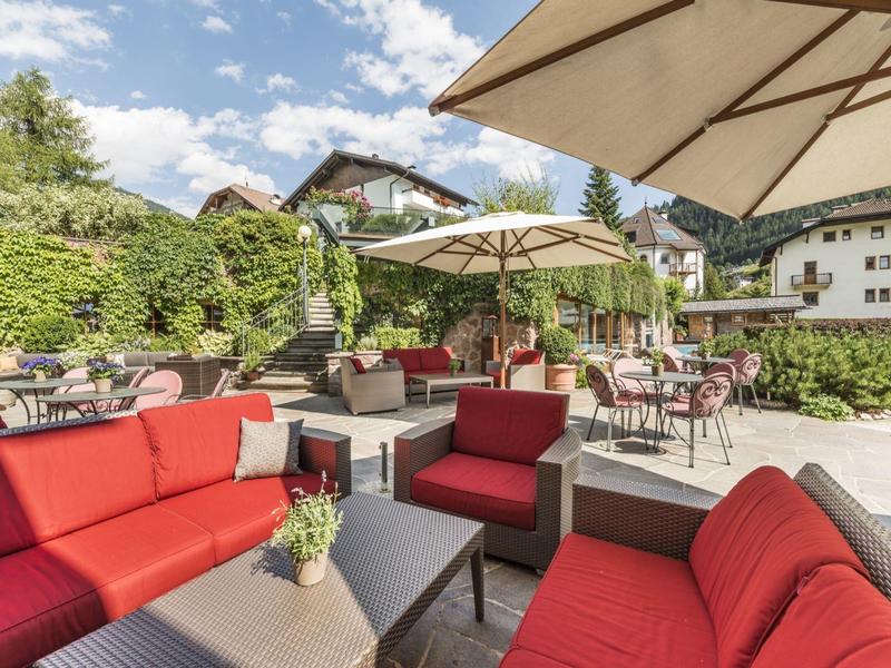 Cozy outdoor terrace with red seating, sun umbrellas, and café tables against a green backdrop.