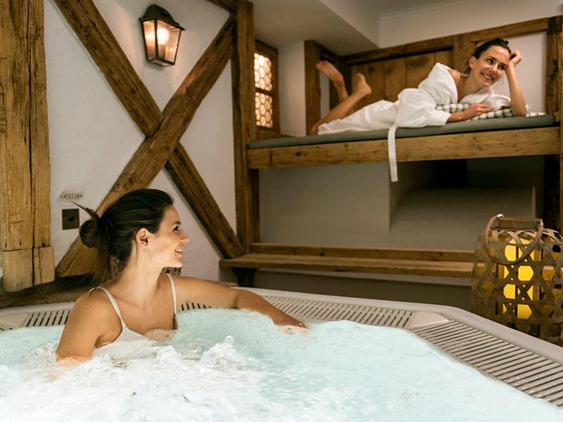 Two women relax in a hot tub and a bunk bed in a warmly lit room.
