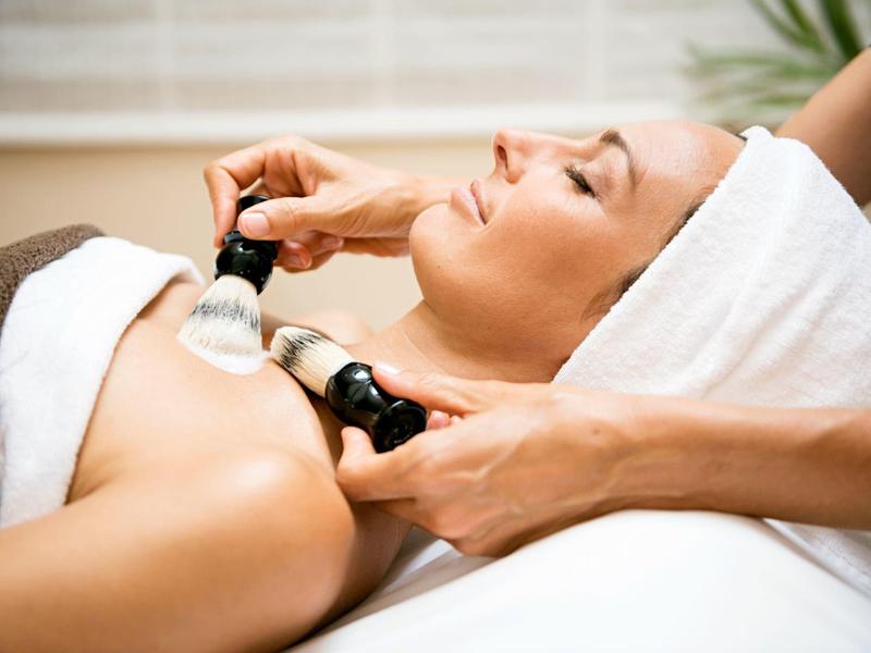 Woman enjoys aesthetic treatment with brushes on décolleté at spa.