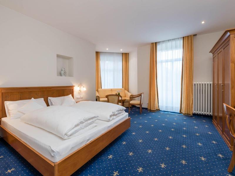 Camera d'hotel luminosa con letto matrimoniale, moquette blu e mobili in legno.