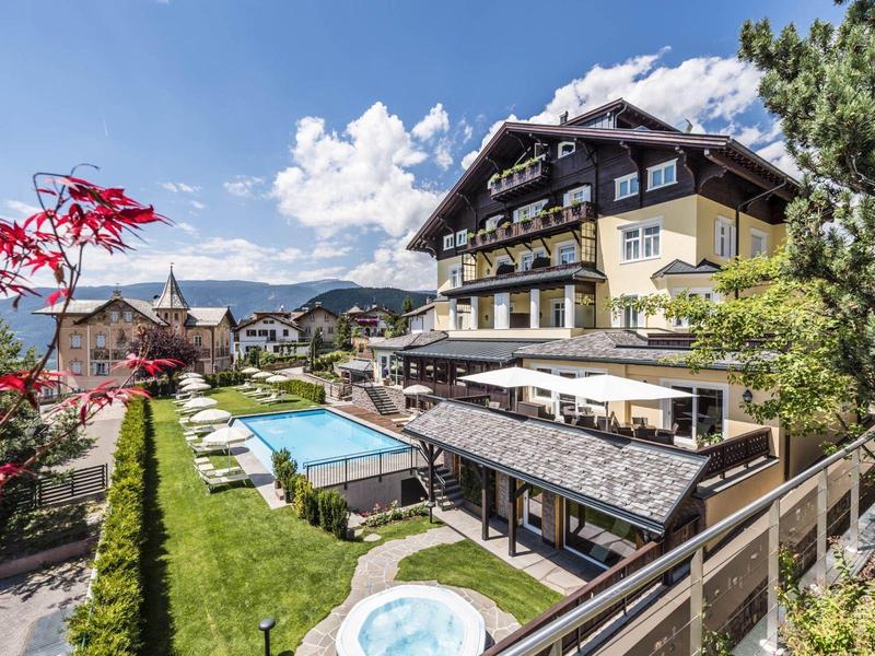 Hotel con piscina, terrazza solarium e montagne sullo sfondo sotto un cielo sereno.