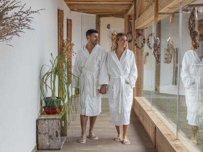 Pár v bílých županech pohodlně prochází chodbou lázní s rostlinami a dekoracemi.