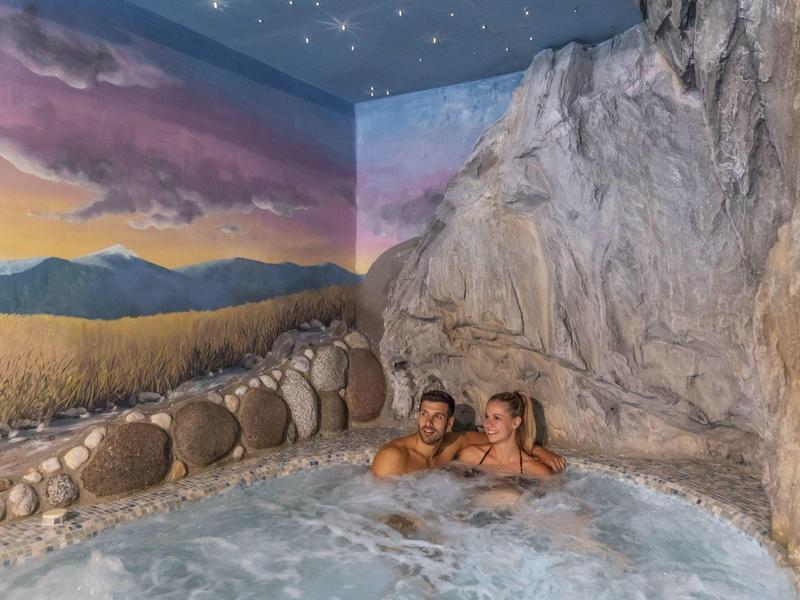 Pár relaxuje ve vířivce s kamennou stěnou a malovanou krajinou nebe.