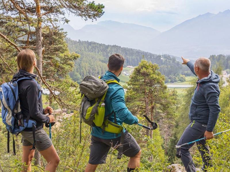 Tři turisté se dívají na horskou zalesněnou krajinu s oblohou a horami v pozadí.