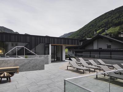 Moderne Dachterrasse mit mehreren Liegestühlen, Holzfeuerstelle, Glasbrüstung und Berge im Hintergrund.