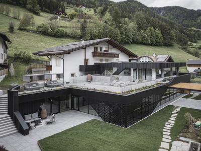 Modernes Haus mit großem Balkon, Terrasse, gepflegtem Rasen und bergiger Landschaft im Hintergrund.