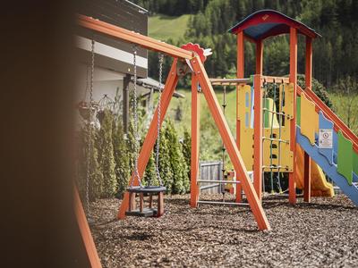 Children's playground with red slide and swing set in hotel garden near wooded hill.