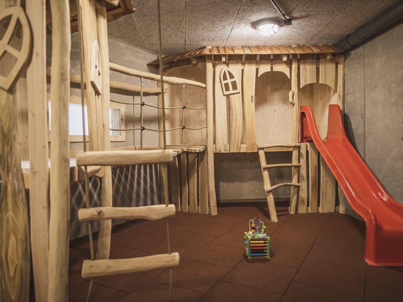 Kinderspielbereich mit Klettergerüst aus Holz und roter Rutsche auf weichem Bodenbelag.