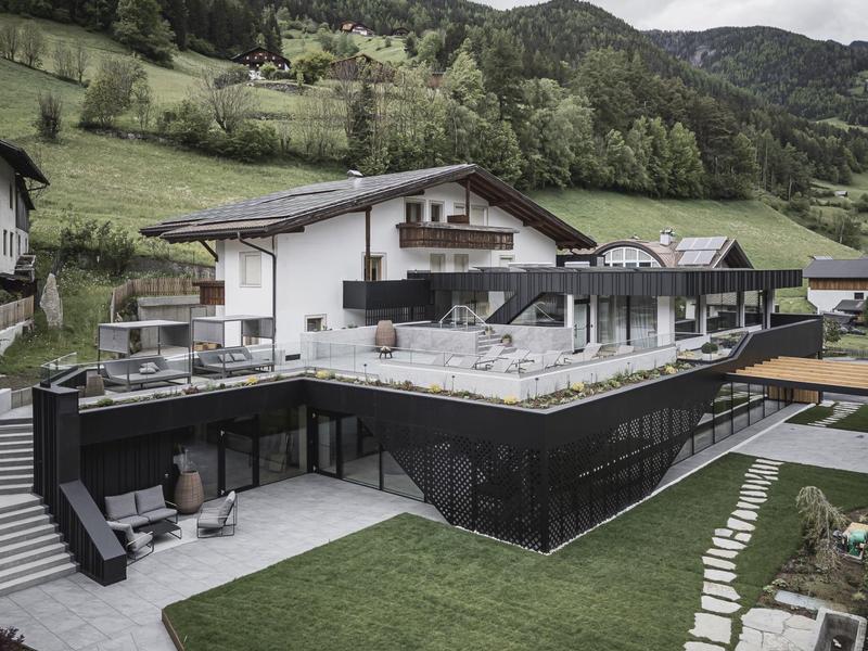 Modernes Haus mit großem Balkon, Terrasse, gepflegtem Rasen und bergiger Landschaft im Hintergrund.