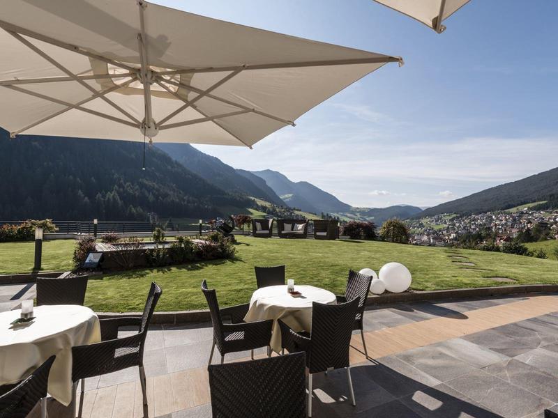 Terrasse mit Tischen, Stühlen und Sonnenschirmen, im Hintergrund Berge und grünes Tal bei Sonnenschein.