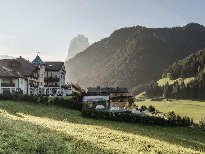 Ein großes weißes Hotel mit Türmen steht auf einer grünen Wiese vor bewaldeten Bergen bei Sonnenaufgang.