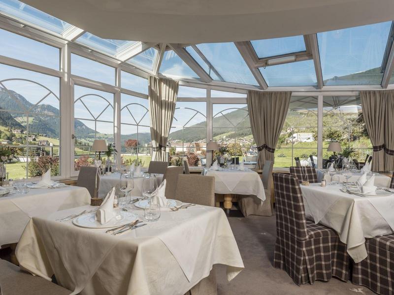 Einladender Wintergarten mit weißen Tischtüchern, karierten Stühlen und Bergblick bei Sonnenschein.