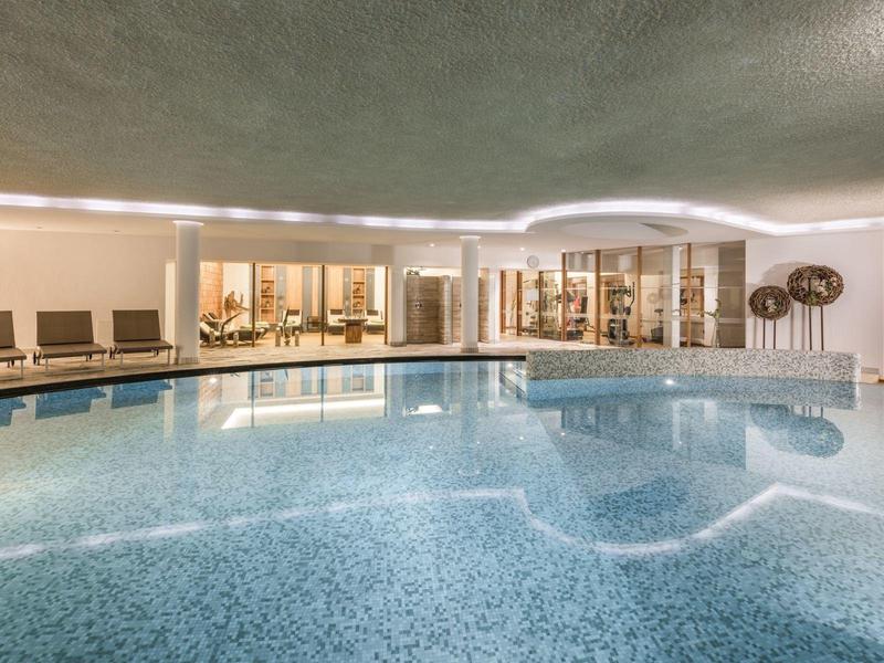 Hell beleuchtetes Hallenbad mit blauem Wasser, Liegestühlen und großen Fenstern zur angrenzenden Lounge.