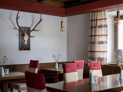 Gemütlicher Gastraum mit Holztischen, roten Sesseln, Gardinen und Hirschgeweih an der Wand.