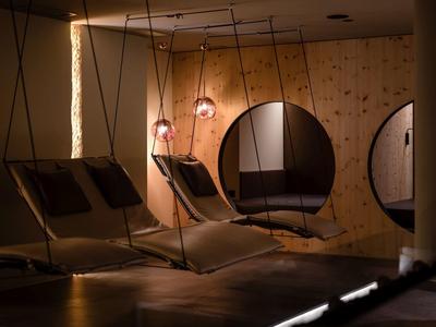 Dunkler Raum mit hängenden Liegen aus Holz und Stoff, warmes Licht und runde Holzfenster an der Wand.