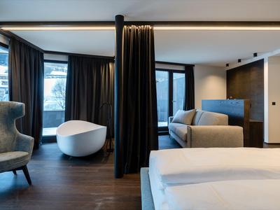 Habitación de hotel moderna con bañera independiente, sofá y cama doble junto a grandes ventanas.