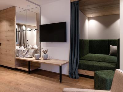 Habitación de hotel moderna con detalles de madera, pared blanca, TV y área de descanso acogedora con tapicería verde oscuro.