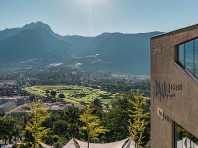 Berge mit Tal, grünen Feldern und Häusern unter strahlender Sonne, rechts modernes Gebäude.