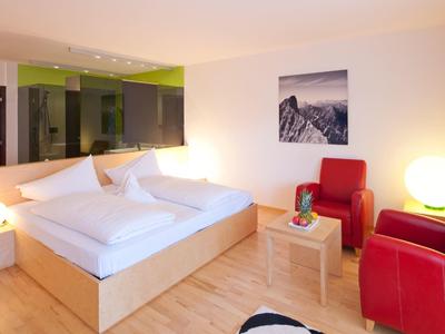 Helles Schlafzimmer mit Doppelbett, roten Sesseln, kleinem Holztisch und Bergbild an der Wand.