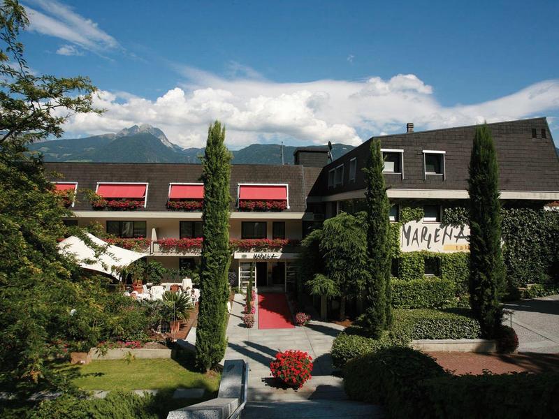 Moderner Gebäudekomplex mit roten Markisen, Zypressen und Bergen im Hintergrund unter blauem Himmel.