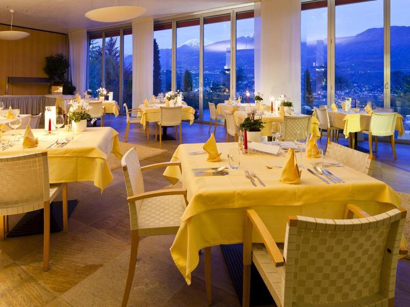 Hell eingerichtetes Restaurant mit gelb gedeckten Tischen und großen Fenstern mit Bergblick.