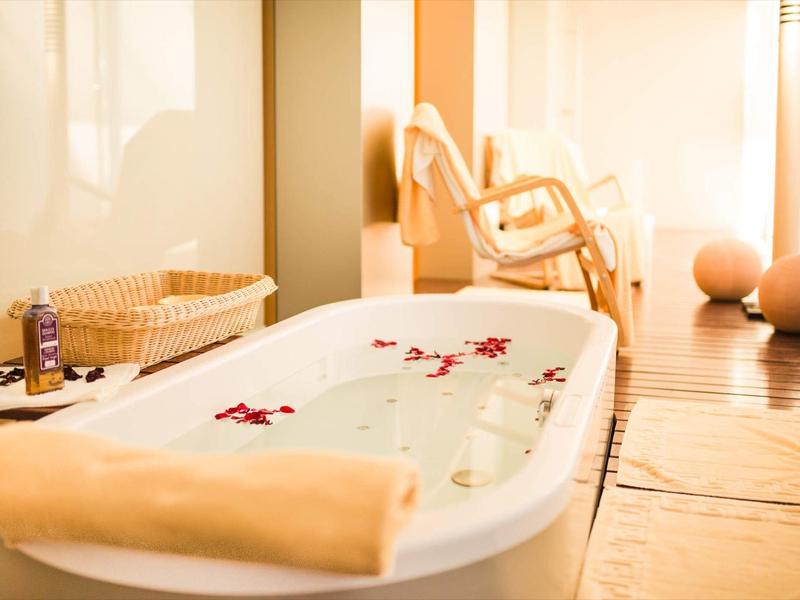 Weißes Bad mit freistehender Badewanne, Holzstuhl, Korb und sanftem, warmem Licht.