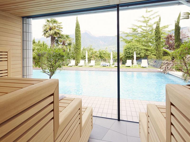 Innenraum einer Sauna mit Holzbank und großer Glasfront zum Außenpool mit Bäumen und Bergen.