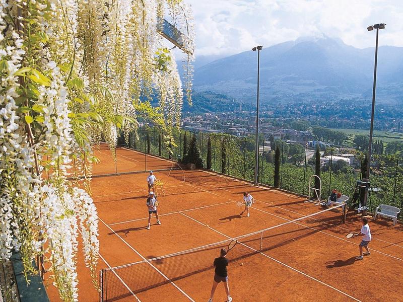 Ein rotes Sandtennisfeld mit Spielern, einer blühenden Kletterpflanze links und Bergen im Hintergrund.