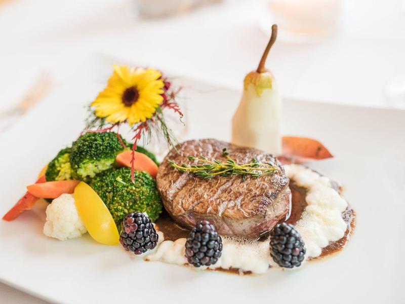 Ein kunstvoll angerichtetes Gericht mit gebratenem Fleisch, Beeren, Gemüse und Blume auf weißem Teller.