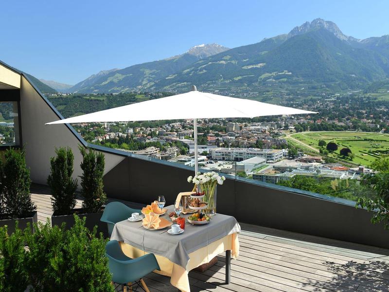 Terrasse mit Tisch, Stühlen, Sonnenschirm und Alpenberg im Hintergrund bei klarem Himmel.