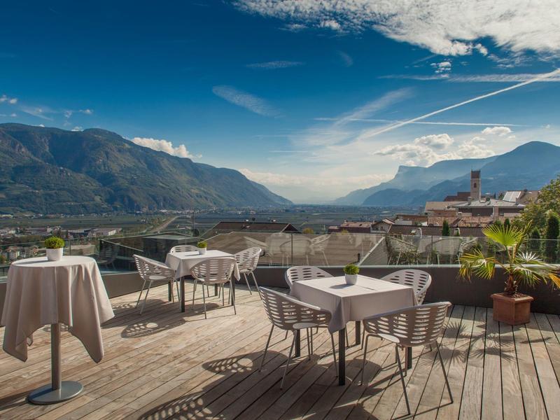 Terrasse mit Tischen und Stühlen, Blick auf Berglandschaft unter blauem Himmel mit Wolken.