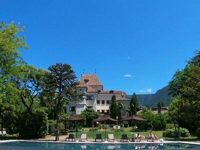 Ein großes Schloss mit roten Dächern steht hinter einem blauen, rechteckigen Pool und grünen Bäumen.