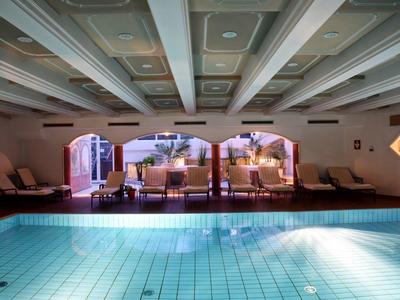 Hallenbad mit blauem Wasser, Sonnenliegen und großen Fenstern mit Blick ins Freie.