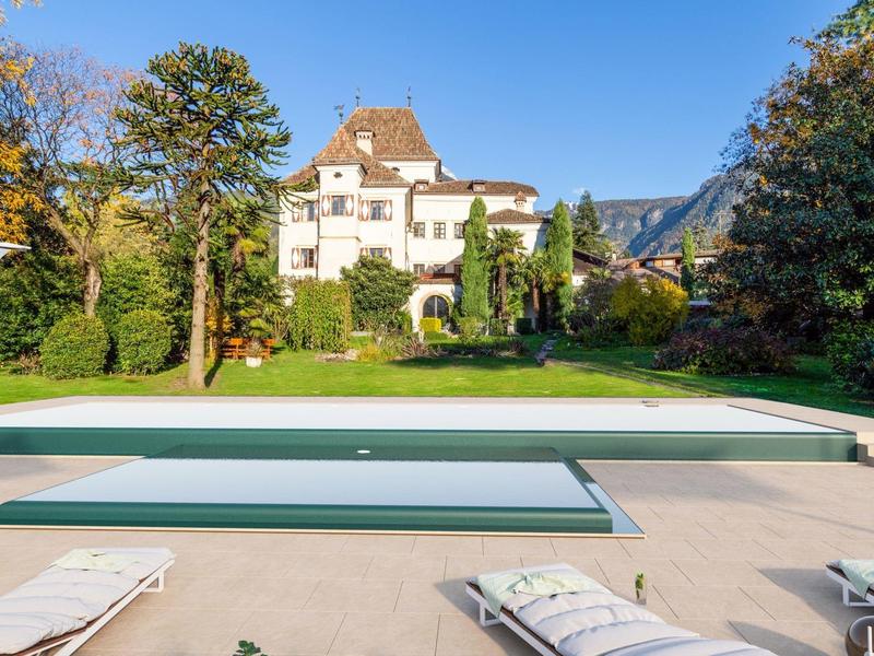 Ein großes Haus in Weiß umgeben von Bäumen und grasgrünem Garten mit Pool und Liegestühlen im Vordergrund.