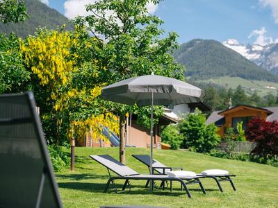 Liegestühle mit Sonnenschirm auf einer grünen Wiese vor Bergen und blühenden Bäumen
